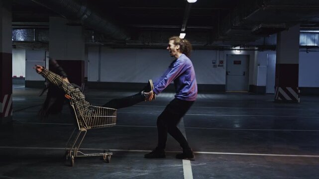 Two lovers act like kids, using a shopping cart as an improvised ride in a parking lot. The guy pushes his girlfriend, both smiling and enjoying a spontaneous, fun moment together.