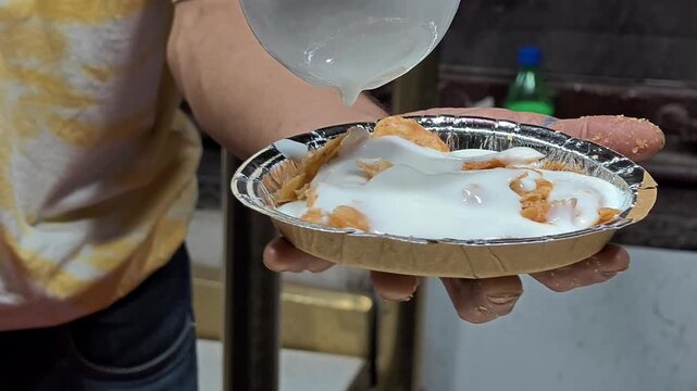 A street food vendor pouring a generous amount of creamy white curd over a plate of crispy fried dough wafers