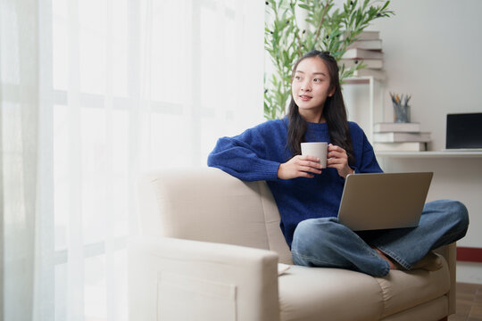 Asian woman working remotely or studying from home, comfortably seated on a sofa with a laptop and coffee, thinking about ideas and projects, enjoying flexible lifestyle remote work balance