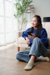 Obraz premium Young Asian woman smiling and holding a smartphone, sitting on the living room floor by a sofa, relaxing while browsing through social media or watching online content at home