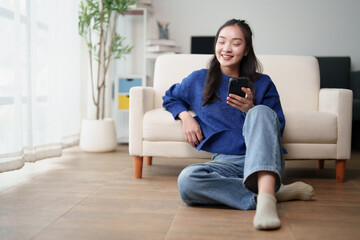 Obraz premium Young asian woman in casual attire relaxing on the floor at home, smiling and looking at her smartphone, engaging in leisure activities and communication