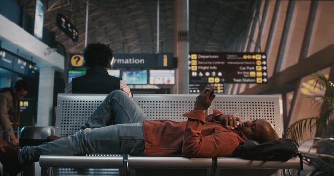 Black Man, Exhausted by Long Layover or Flight Delay, Lies Across Metal Seats in Airport Departure Lounge. Holding Smartphone Above Face, Male Scrolls Through Content Pass Time. Exhaustion of Transit.