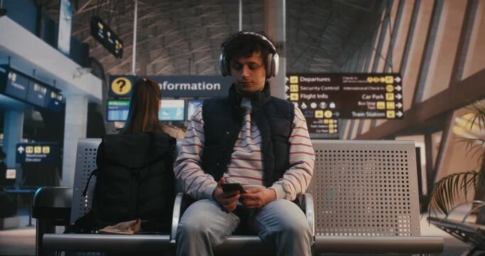 Young Man Sits in Airport Terminal, Wearing Large Silver Over Ear Headphones. Guy Listens Music While Scrolling Through Smartphone and Eventually Lifts Device to Take Selfie or Record Video Message.