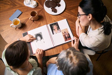 High angle view of women reviewing family photo album at table, pointing at portraits and sharing memories. Suitable for genealogy, family history, memory care, legacy planning marketing