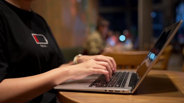 Close-up of person hands typing on laptop keyboard in cafe at night, female freelancer working on computer with bokeh background, digital nomad and remote work concept.