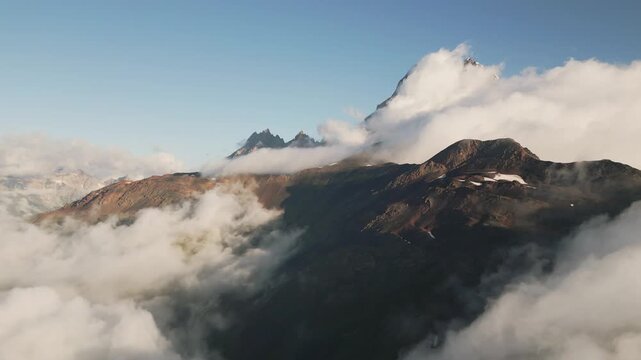 Aerial view of Mount Ushba peak partially covered in morning clouds. A dramatic mountain landscape in Svaneti, Georgia, featuring rugged peaks and atmospheric mist in the Caucasus.