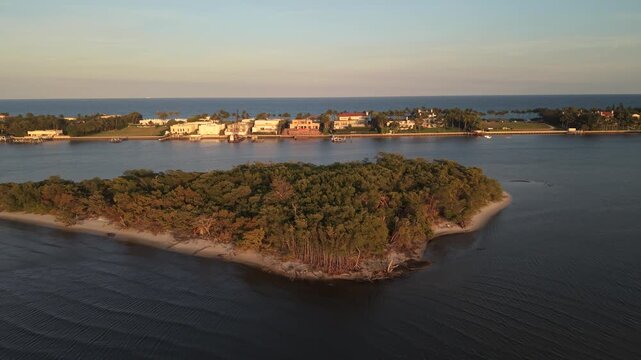 Drone in golden hour flying east over the ICW, from Summa Beach Park in West Palm Beach Florida toward the barrier island showing waterfront homes and the Atlantic Ocean in the distance.