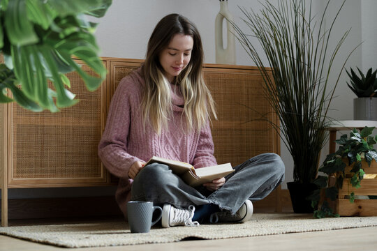 Young woma reading a book on rug at home surrounded by plants