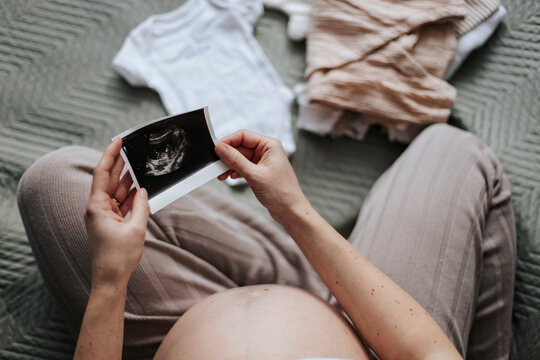 Pregnant woman holding ultrasound scan with newborn clothing at home