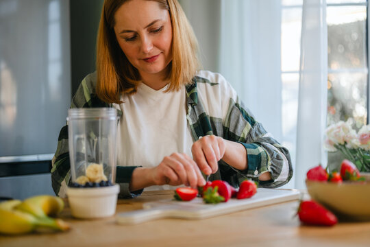 Woman reparing fresh berry smoothie with banana in home kitchen