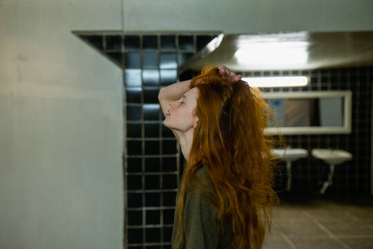Red-haired woman with freckles in old tiled restroom touching hair
