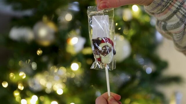 Gros plan d'un enfant ouvrant une sucette en chocolat en forme de t&ecirc;te de p&egrave;re no&euml;l, sapin de no&euml;l avec guirlande lumineuse en fond.