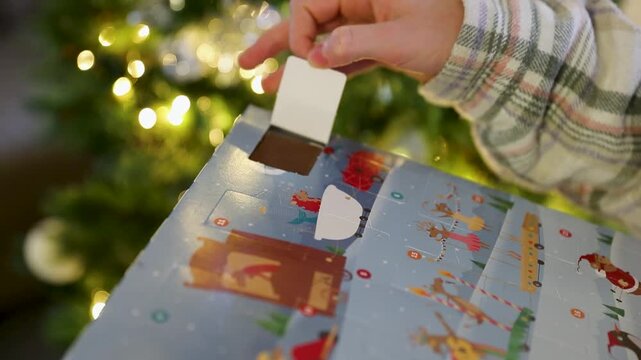 Gros plan d'un enfant ouvrant la fen&ecirc;tre d'un calendrier de l'Avent avec un chocolat, sapin de no&euml;l avec guirlande lumineuse en fond.