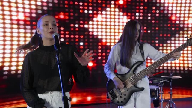 A female music band performs on stage. The lead singer is at the microphone while a bassist plays her guitar. A bright, colorful LED screen is in the background. They are dressed in black and white