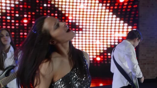 A female singer in a sparkly dress energetically performs with her music band on stage. Guitarists and a drummer play in front of a bright, flashing red and white LED screen