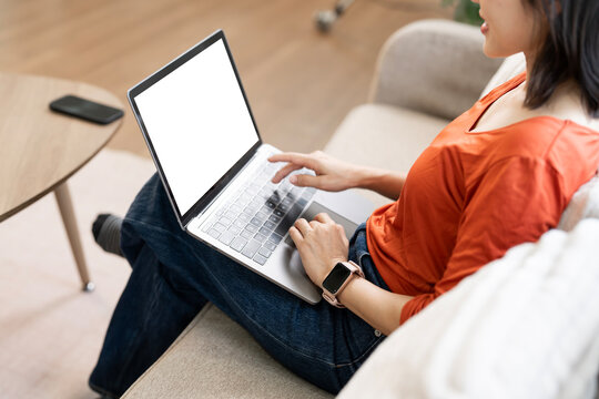 Young female freelancer sitting on sofa and using laptop with blank white mockup screen. Work from home concept.