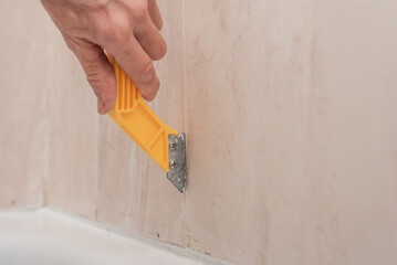 Closeup of male plumber removing old silicone in bathroom with special tool. Removing old grout between bathroom tiles with a special scraper