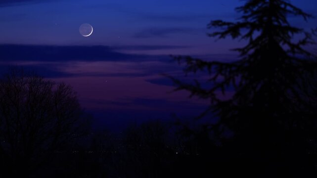 Blue hour time with crescent Moon, stars and planets above landscape silhouettes.