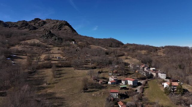 Monte Lama with San Biagio hamlet in Northern Italy, featuring rocky ridge, scattered rural houses and hillside landscape with dry vegetation and trees, aerial glide shot.
