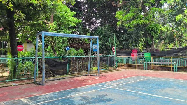 Still shot of an empty outdoor futsal and basketball court with a metal goal post surrounded by lush green trees