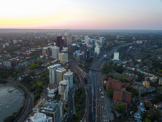 Fototapeta premium Aerial drone view of Sydney city skyline featuring modern office buildings and busy highway traffic in North Sydney during sunset. The image captures urban infrastructure, transportation, and resident