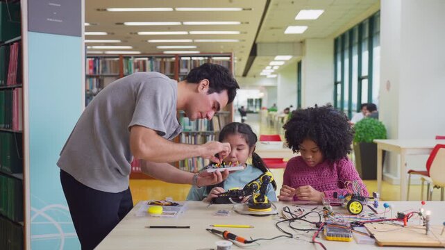 Teacher assists two young girls working on robotics project in library. 