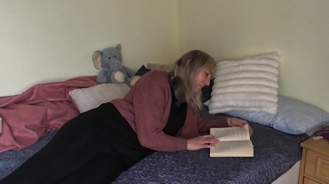 Mature woman lying on bed reading book and searching for information at home