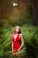 Naklejka premium A young woman in a red dress stands among tall green ferns in a dense forest.