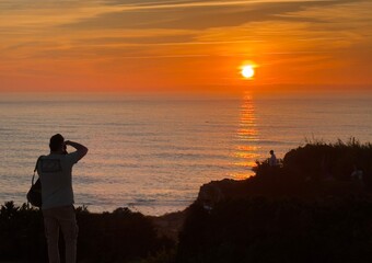 Fototapeta premium Photographer silhouetted on a cliff documenting a vibrant golden hour sunset over the sea