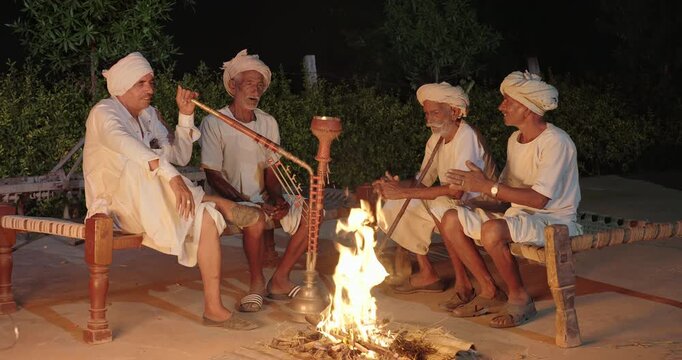 Indian old aged man wear white kurta pajama sitting on woven cot bed enjoy desi life at rural area. Happy four male group talk speak share gossip do hookah hold pipe spend night time village place