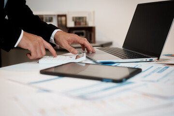 Fototapeta premium Businessperson using calculator and laptop on desk with tablet and financial charts, office background, close-up hands doing accounting and budget analysis.