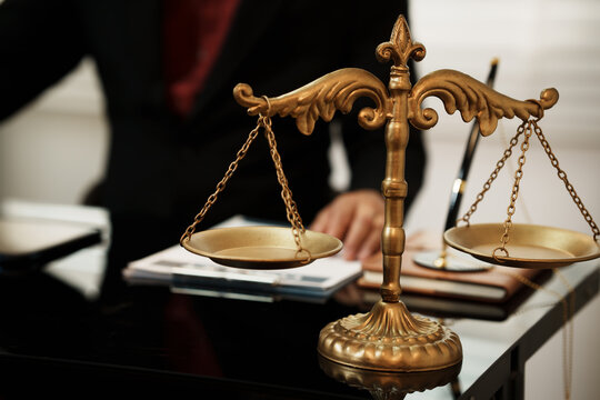 Close-up of brass scales of justice on desk with blurred lawyer signing documents, legal office concept, shallow depth of field.
