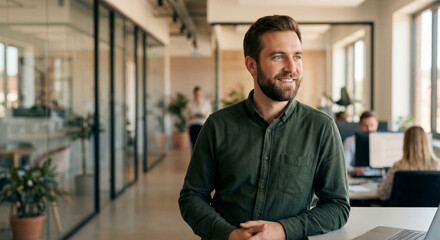 Obraz premium Handsome and confident male professional in a green shirt smiles and looks aside in a bright, modern open-plan office