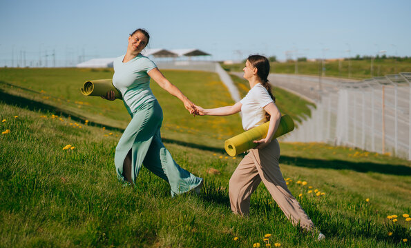 Two women joyfully ascending a grassy hill under a clear blue sky, each holding a yoga mat, symbolizing friendship and connection with nature.