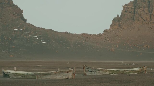 Two decayed wooden whale boats lie abandoned on volcanic black sand at Whalers Bay, Deception Island, as tourists hike rocky ridge trail between dramatic cliffs in background