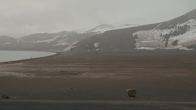 Wide panoramic view of vast dark volcanic ash plain curving alongside calm caldera waters at Whalers Bay, Deception Island, with snow-draped volcanic slopes fading into haze