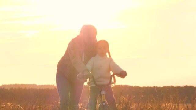 Mom teaching child to ride bike, silhouettes at sunset in field. Caring, careful, gentle and loving approach to teaching instructing training girl to ride bike. Touching shots of happy joyful family.