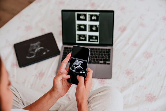 Pregnant woman viewing ultrasound on phone at home with laptop