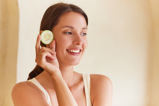 Cucumber eye treatment woman holding a fresh cucumber slice near her eye with a wide smile, looking sideways. Natural skincare concept, beige background, large copy space right.