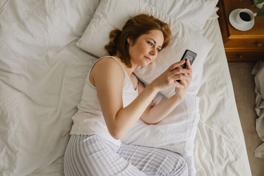 Person lying in bed scrolling on smartphone in morning bedroom