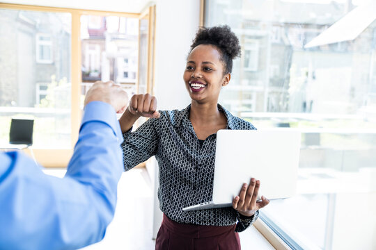 Smiling colleague giving fist bump holding laptop in modern office