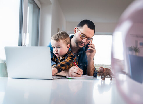 Father multitasking in home office with son and notebook