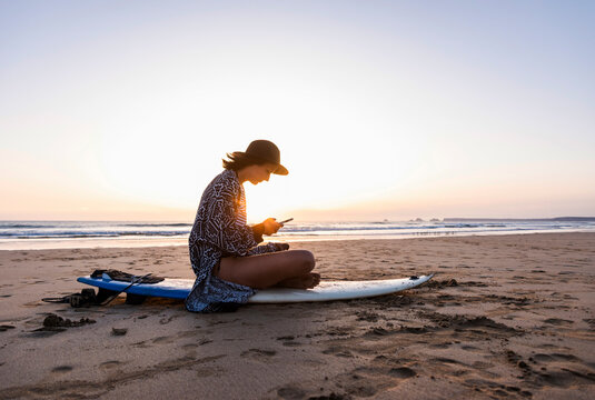 Woman sitting on surfboard at the beach messaging on mobile at sunset