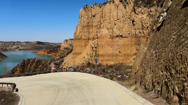 High angle drone shot showing a winding mountain road alongside a tranquil reservoir at the base of steep sediment cliffs and arid canyon terrain.
