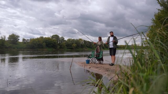 Young caucasian man and woman spending their leisure time fishing together with rods from a sandy river bank on a cloudy day, enjoying their outdoor hobby and relaxing in nature