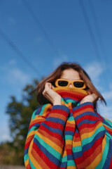 Fototapeta premium Young woman in a rainbow striped sweater and yellow sunglasses outdoors, playful pose pulling sweater over her lower face, casual outfit with blue sky background for lifestyle travel content.