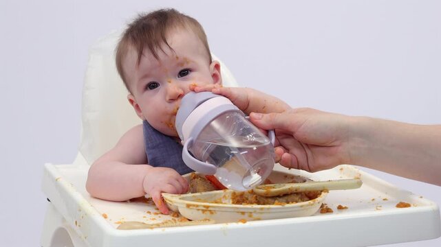 White background. A little girl in a high chair, picking and eating food from a sectioned plate with her fingers. The concept of weaning is based on the principle of "child-choosing food