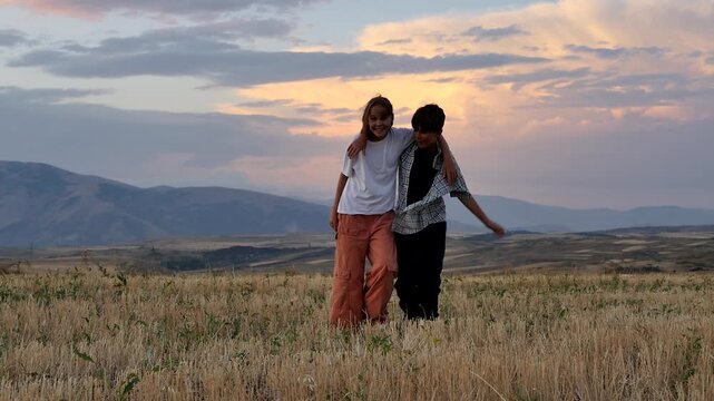 Teen boy and girl high five and walk together hugging across mowed rural field in mountain area at sunset. Lifelong friends. Brother and sister strong bond. Warm family relationships