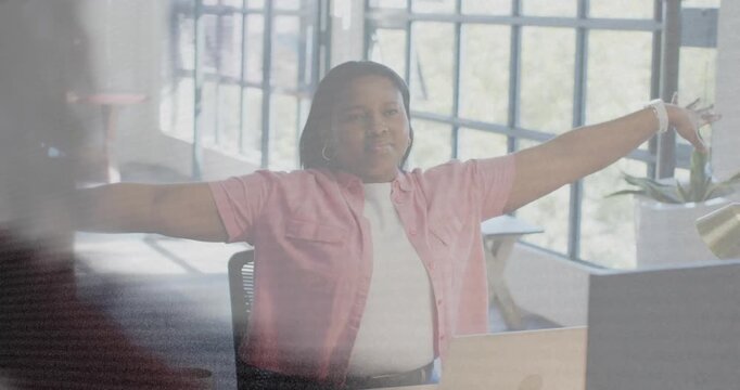 Woman stretching then responding to blurred colleague at desk with laptop in business office