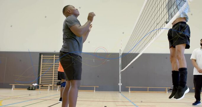 Tall man in gray tee stepping to net, holding volleyball with teammate moving and jumping, training